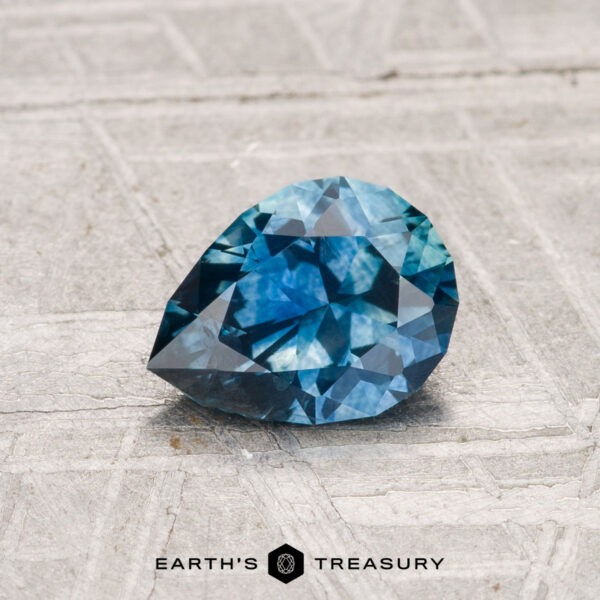 1.04-Carat Montana Sapphire (Heated) A 1.04-carat heated Montana sapphire, faceted in a pear shape, rests on a light gray textured surface. The "EARTH'S TREASURY" logo is displayed at the bottom of the image.