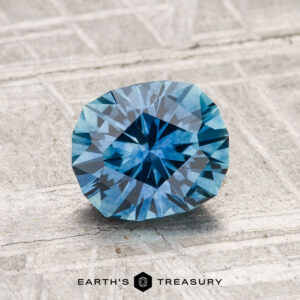 A 1.42-carat heated Montana sapphire with a faceted geometric pattern rests on a textured light gray surface. "EARTH'S TREASURY" is shown at the bottom.
