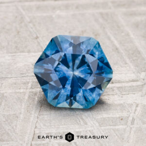 A 1.46-carat heated Montana Sapphire with a hexagonal blue cut and intricate facets rests on a textured gray surface. The "EARTH'S TREASURY" logo and text are visible at the bottom of the image.