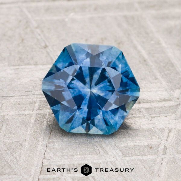 A 1.46-carat heated Montana Sapphire with a hexagonal blue cut and intricate facets rests on a textured gray surface. The "EARTH'S TREASURY" logo and text are visible at the bottom of the image.