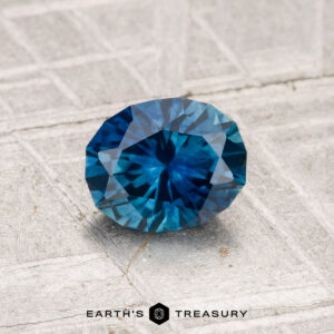 A faceted oval 1.00-carat heated Montana Sapphire rests on a textured, light gray surface with the text "EARTH'S TREASURY" below.