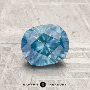 A 1.39-carat heated Montana sapphire with blue facets rests on a textured gray surface, with "EARTH'S TREASURY" displayed below.