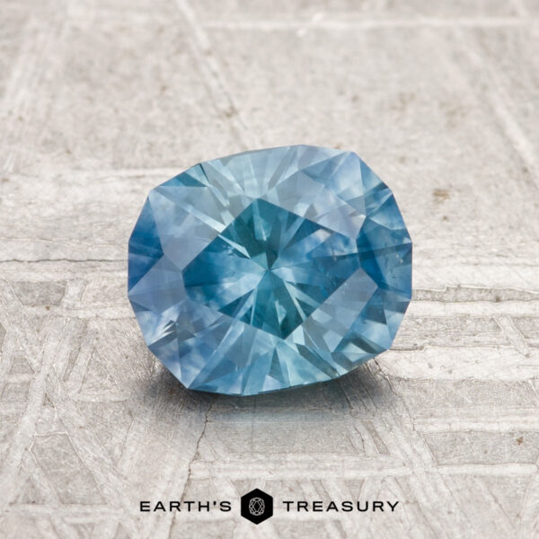 1.39-Carat Montana Sapphire (Heated) A 1.39-carat heated Montana sapphire with blue facets rests on a textured gray surface, with "EARTH'S TREASURY" displayed below.