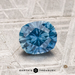 A 1.38-carat heated Montana sapphire with a faceted blue appearance rests on a textured gray surface, displaying the "Earth's Treasury" logo and text at the bottom.