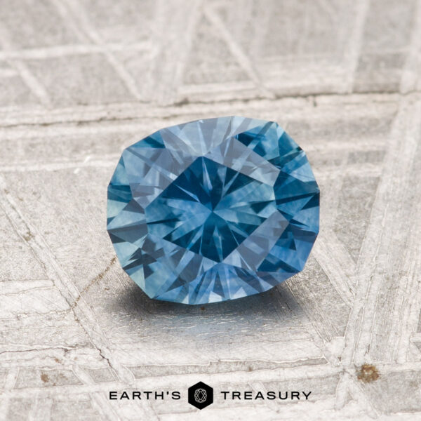 1.38-Carat Montana Sapphire (Heated) A 1.38-carat heated Montana sapphire with a faceted blue appearance rests on a textured gray surface, displaying the "Earth's Treasury" logo and text at the bottom.