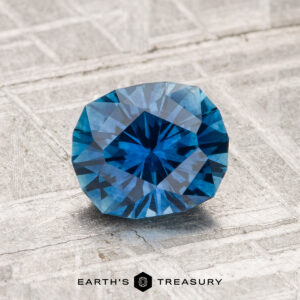 A 1.38-carat heated Montana sapphire with a faceted blue appearance displayed on a textured gray surface, featuring the text "EARTH'S TREASURY" at the bottom.