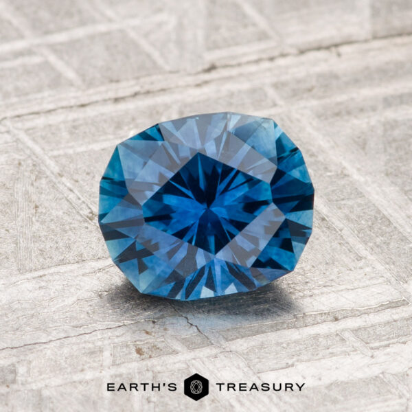 1.38-Carat Montana Sapphire (Heated) A 1.38-carat heated Montana sapphire with a faceted blue appearance displayed on a textured gray surface, featuring the text "EARTH'S TREASURY" at the bottom.