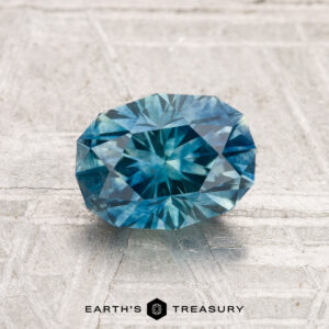 A faceted 1.62-carat heated Montana sapphire with intricate cuts rests on a textured light surface; the "EARTH'S TREASURY" logo appears at the bottom.