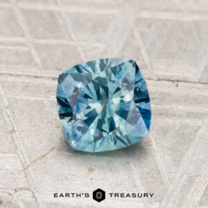 A cushion-cut 1.08-carat heated Montana sapphire displayed on a textured light gray surface with "EARTH'S TREASURY" below.