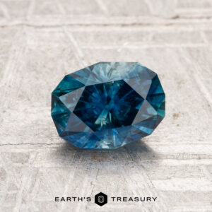 A 1.36-carat heated Montana sapphire, oval faceted and blue, rests on a light textured surface with "EARTH'S TREASURY" at the bottom.