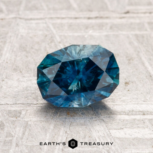 1.36-Carat Montana Sapphire (Heated) A 1.36-carat heated Montana sapphire, oval faceted and blue, rests on a light textured surface with "EARTH'S TREASURY" at the bottom.