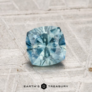A 1.01-carat heated Montana sapphire with a cushion cut is displayed on a light textured surface, with "Earth's Treasury" written below the gemstone.