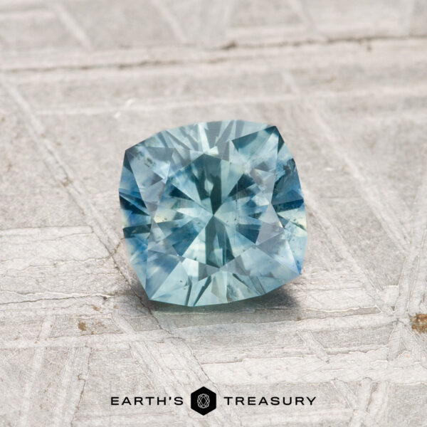 1.01-Carat Montana Sapphire (Heated) A 1.01-carat heated Montana sapphire with a cushion cut is displayed on a light textured surface, with "Earth's Treasury" written below the gemstone.