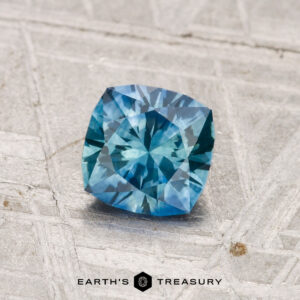 A 0.96-carat heated Montana sapphire, cushion-cut with intricate facets, rests on a textured gray surface. The "EARTH'S TREASURY" logo appears at the bottom.