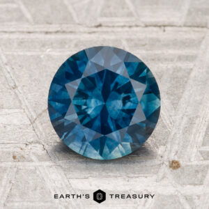 A 1.78-carat heated Montana sapphire, round and faceted, rests on a textured light gray surface with "Earth's Treasury" displayed at the bottom.