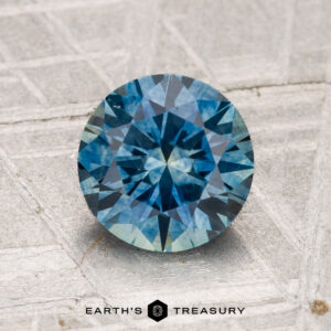 A 1.77-carat heated Montana sapphire, round brilliant-cut and blue, displayed on a textured light surface with "Earth's Treasury" at the bottom.