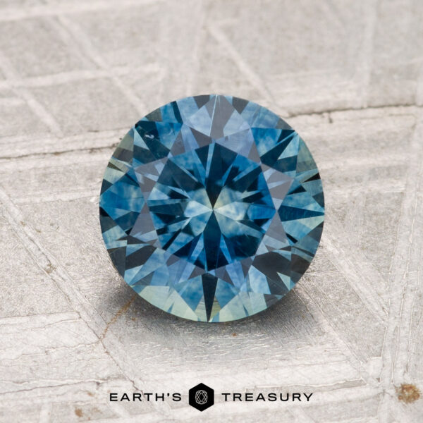1.77-Carat Montana Sapphire (Heated) A 1.77-carat heated Montana sapphire, round brilliant-cut and blue, displayed on a textured light surface with "Earth's Treasury" at the bottom.