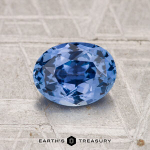A 1.62-carat Ceylon Sapphire, faceted and oval-shaped, rests on a patterned metallic surface with "EARTH'S TREASURY" displayed below.