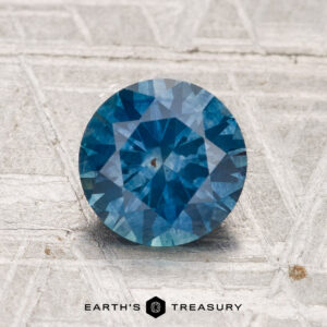 A round, faceted 1.63-carat heated Montana sapphire is displayed on a textured, light-colored surface, with the text "EARTH'S TREASURY" shown below.
