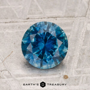A 1.60-carat heated Montana sapphire, round brilliant-cut and blue, is displayed on a textured light gray surface with "EARTH’S TREASURY" at the bottom.