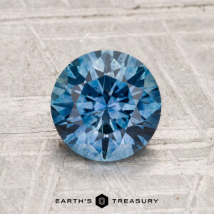 A 1.57-carat heated Montana Sapphire in a round brilliant cut is displayed on a light textured surface with the text "Earth's Treasury" below it.