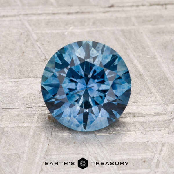 1.57-Carat Montana Sapphire (Heated) A 1.57-carat heated Montana Sapphire in a round brilliant cut is displayed on a light textured surface with the text "Earth's Treasury" below it.