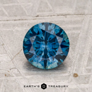 1.56-carat heated Montana Sapphire, round brilliant-cut, showcased on a textured gray surface with "EARTH'S TREASURY" at the bottom.