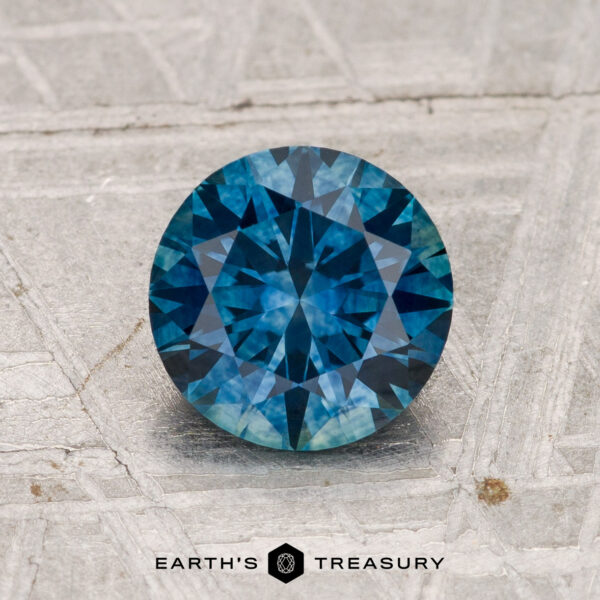 1.56-Carat Montana Sapphire (Heated) 1.56-carat heated Montana Sapphire, round brilliant-cut, showcased on a textured gray surface with "EARTH'S TREASURY" at the bottom.