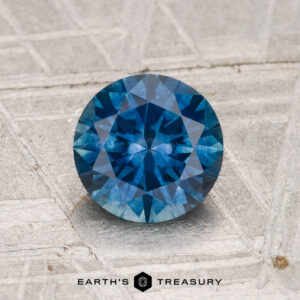 A 1.52-carat heated Montana sapphire with a round brilliant cut is shown on a textured gray background, featuring the text "EARTH'S TREASURY" at the bottom.
