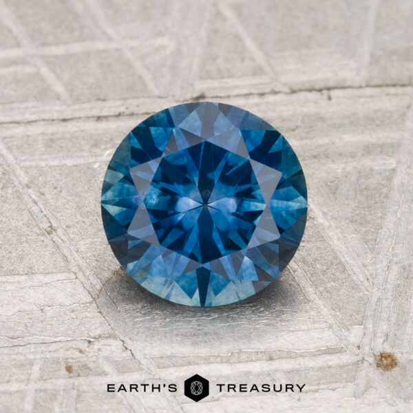 1.52-Carat Montana Sapphire (Heated) A 1.52-carat heated Montana sapphire with a round brilliant cut is shown on a textured gray background, featuring the text "EARTH'S TREASURY" at the bottom.
