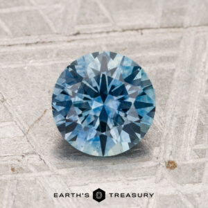 A 1.52-carat heated Montana sapphire, round brilliant-cut and blue, rests on a textured gray surface with "EARTH'S TREASURY" at the bottom.