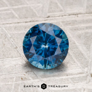 A 1.50-carat heated Montana Sapphire, round brilliant-cut and blue, is displayed on a textured silver-gray surface with the Earth's Treasury logo below.