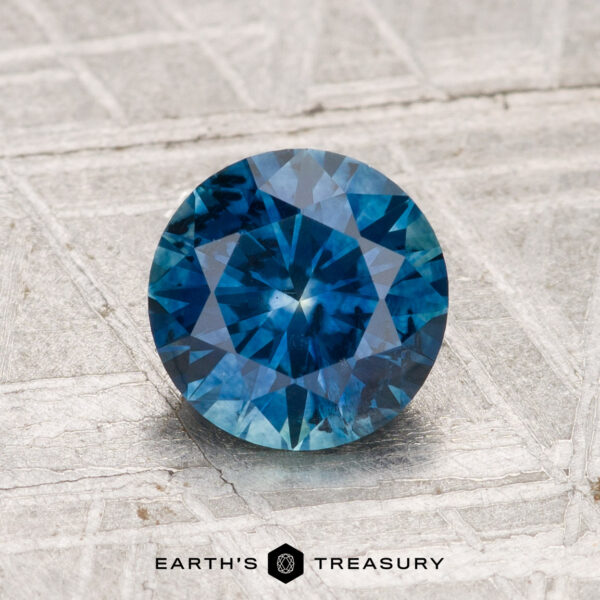 1.50-Carat Montana Sapphire (Heated) A 1.50-carat heated Montana Sapphire, round brilliant-cut and blue, is displayed on a textured silver-gray surface with the Earth's Treasury logo below.