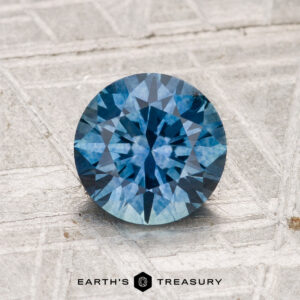 A 1.48-carat heated Montana sapphire with a round brilliant cut is displayed on a textured, metallic surface. "EARTH'S TREASURY" appears at the bottom of the image.