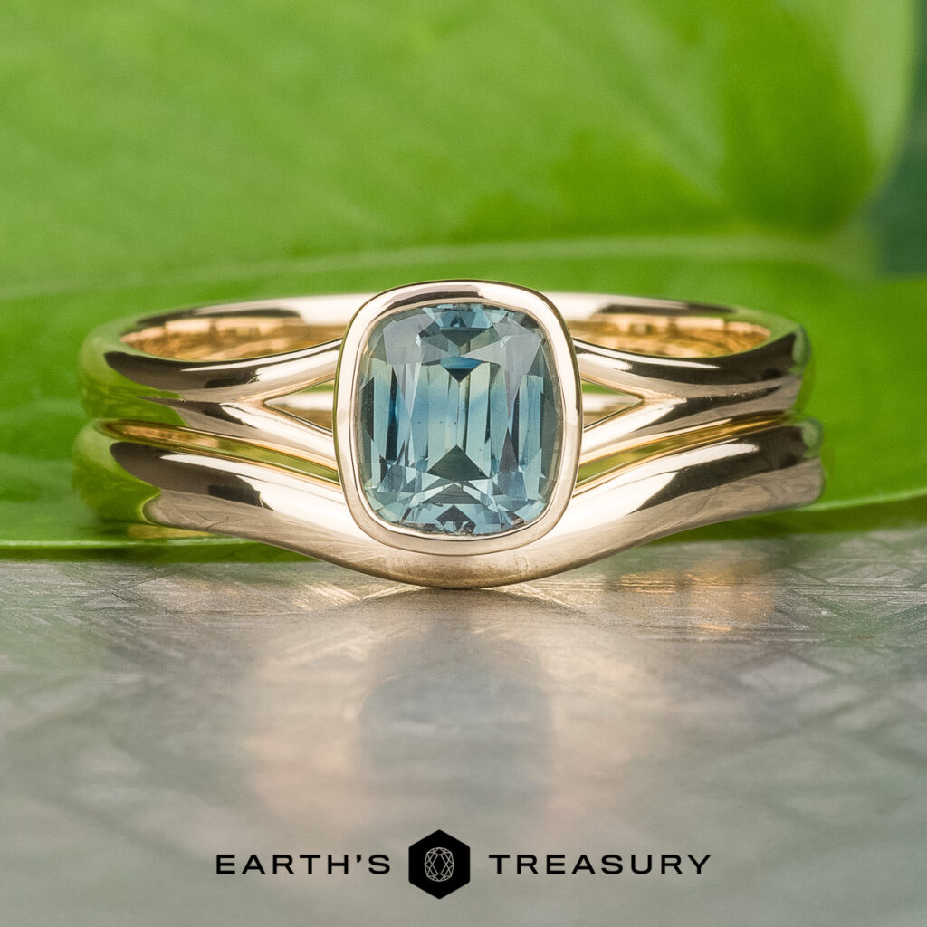 The _SNY0074-Edit-1200-BW ring, featuring a rectangular blue gemstone set in gold, is showcased on a green leaf. The "EARTH'S TREASURY" logo and text appear at the bottom of the image.