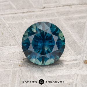 A 1.47-carat heated Montana sapphire, round and faceted in teal, is displayed on a textured gray surface with "EARTH'S TREASURY" at the bottom.