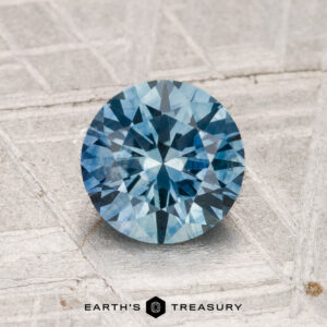 A 1.46-carat heated Montana sapphire with a round brilliant cut rests on a textured metallic surface, "EARTH'S TREASURY" displayed below.