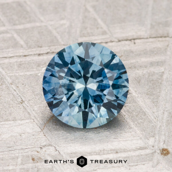 1.46-Carat Montana Sapphire (Heated) A 1.46-carat heated Montana sapphire with a round brilliant cut rests on a textured metallic surface, "EARTH'S TREASURY" displayed below.