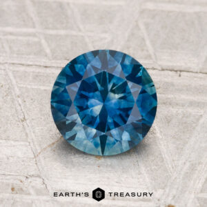 A 1.45-carat heated Montana sapphire, round brilliant-cut and blue, is displayed on a textured gray surface with "EARTH'S TREASURY" at the bottom.