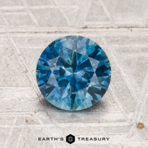 1.44-Carat heated Montana Sapphire, round brilliant-cut blue gemstone on textured silver surface, with "Earth's Treasury" logo and text below.