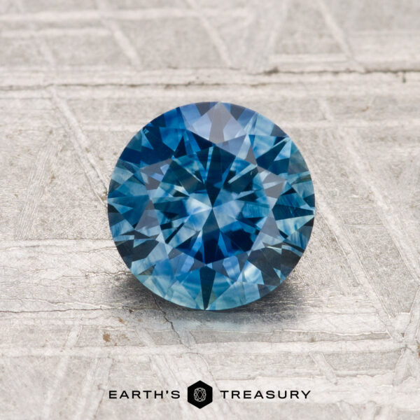 1.42-Carat Montana Sapphire (Heated) A 1.42-carat heated Montana sapphire, round brilliant-cut and blue in color, rests on a textured metallic surface with "EARTH'S TREASURY" displayed at the bottom.