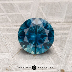 A 1.42-carat round-cut heated Montana sapphire with a deep blue hue is shown on a textured gray surface, featuring the "EARTH'S TREASURY" text and logo at the bottom.