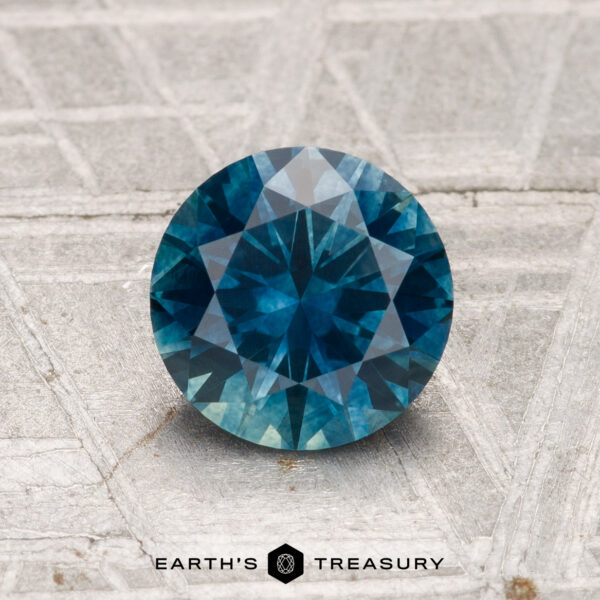 1.42-Carat Montana Sapphire (Heated) A 1.42-carat round-cut heated Montana sapphire with a deep blue hue is shown on a textured gray surface, featuring the "EARTH'S TREASURY" text and logo at the bottom.