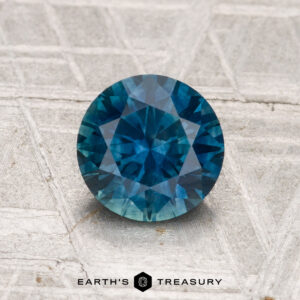 A 1.39-carat heated Montana sapphire, round and faceted, is displayed on a textured gray surface with "EARTH'S TREASURY" shown below.
