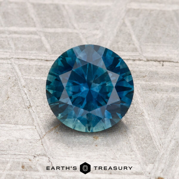 1.39-Carat Montana Sapphire (Heated) A 1.39-carat heated Montana sapphire, round and faceted, is displayed on a textured gray surface with "EARTH'S TREASURY" shown below.