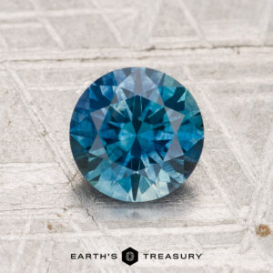 A 1.37-carat heated Montana sapphire, round brilliant-cut and blue, rests on a textured gray surface with "Earth's Treasury" displayed below.