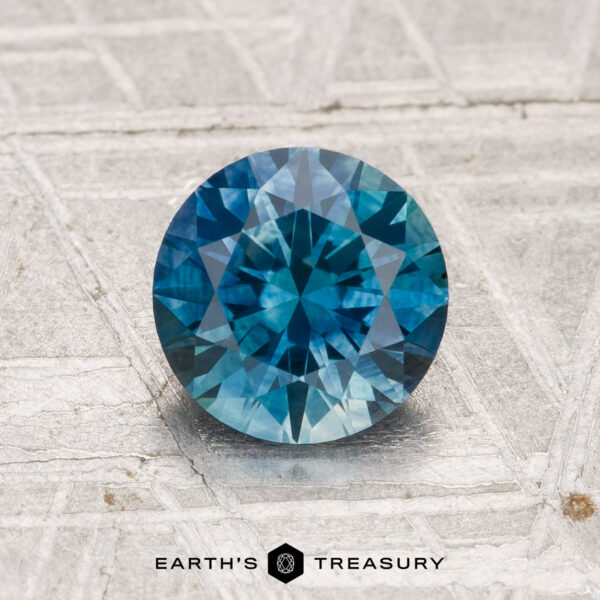 1.37-Carat Montana Sapphire (Heated) A 1.37-carat heated Montana sapphire, round brilliant-cut and blue, rests on a textured gray surface with "Earth's Treasury" displayed below.