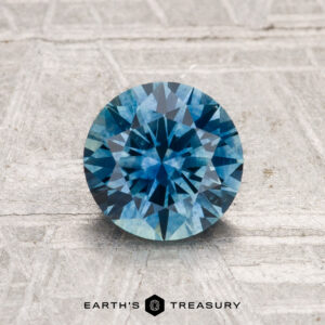 A 1.35-carat heated Montana Sapphire, round brilliant-cut and blue, is displayed on a textured gray surface with "Earth's Treasury" at the bottom.