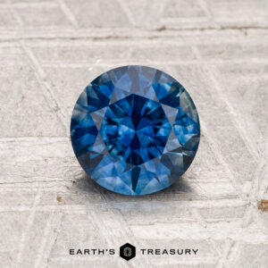 A 1.35-carat heated Montana sapphire with a round brilliant cut rests on a textured light gray surface, with "EARTH'S TREASURY" displayed below.
