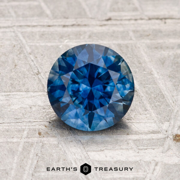 1.35-Carat Montana Sapphire (Heated) A 1.35-carat heated Montana sapphire with a round brilliant cut rests on a textured light gray surface, with "EARTH'S TREASURY" displayed below.
